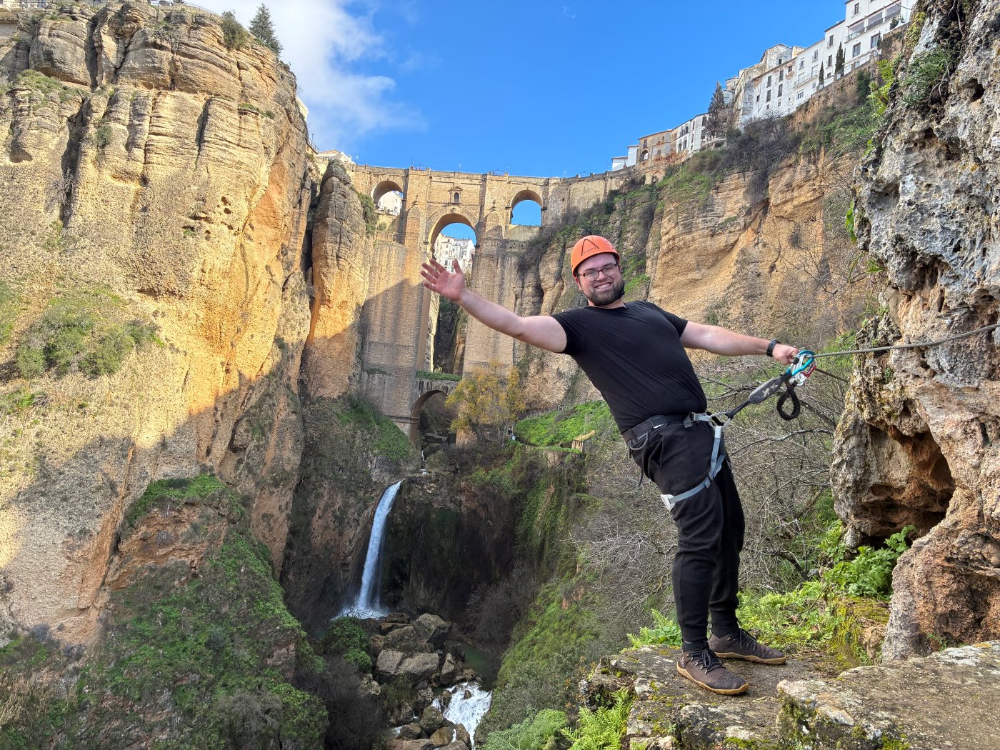 Dan on a via ferrata route partway down a gorge, one arm flung out, a stone arch bridge and waterfall behind him