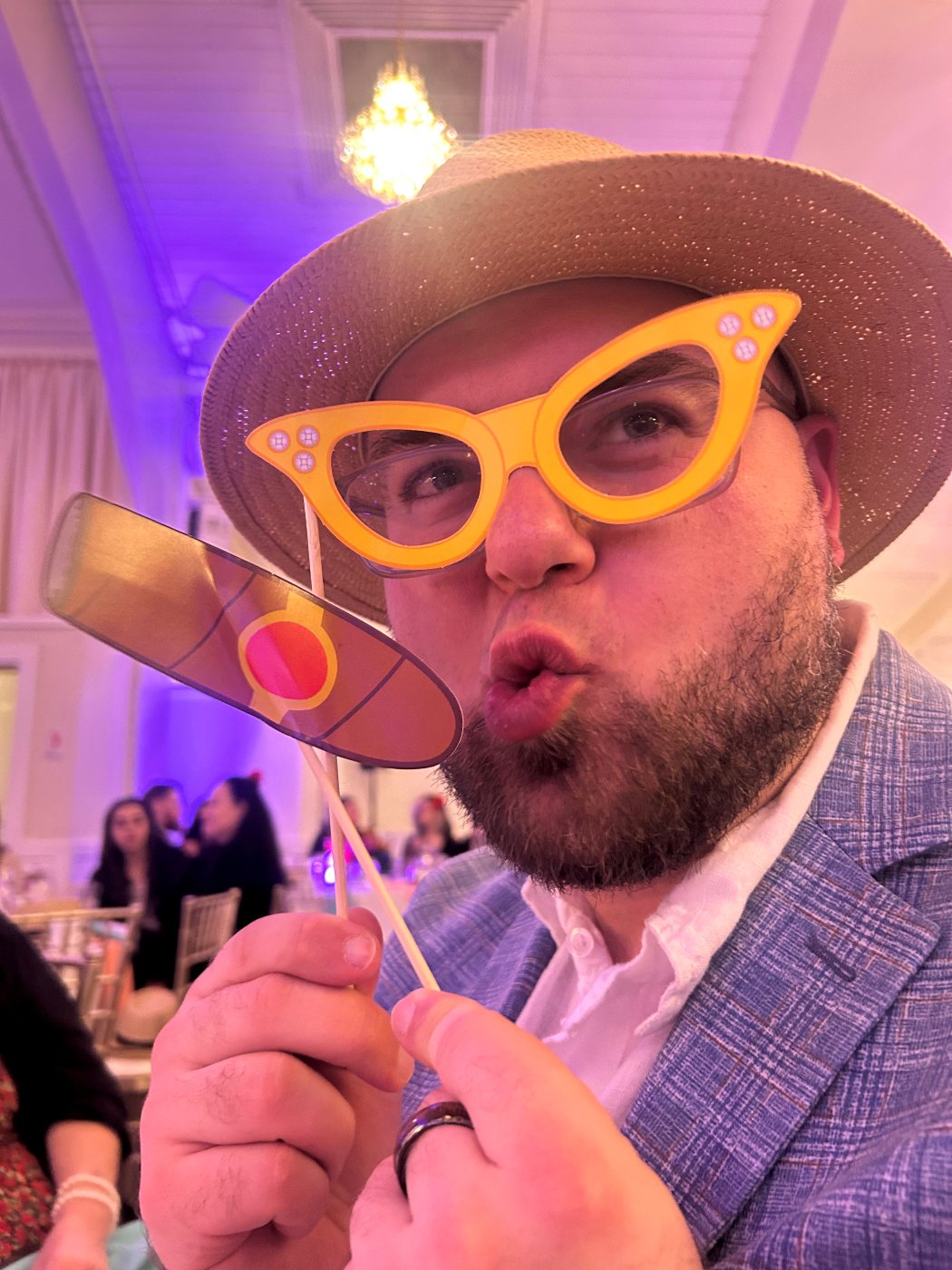 Dan at a wedding in a straw hat, holding oversized novelty glasses and a cigar cutout on sticks in front of his face
