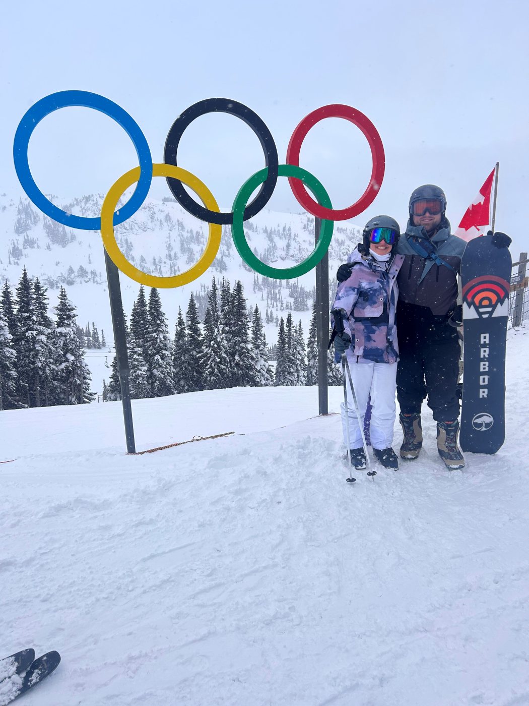 Dan in snowboarding gear holding his board beside a set of Olympic rings, on a snowy mountain in falling snow